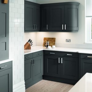 Anthracite shaker units create a modern twist in this traditional kitchen design. White worktops and chrome handles are displayed throughput the kitchen. This viewpoint shows us the kitchen from across the island. White brick is shown on the walls and a light wooden floor. The corner of the kitchen run has a knife set , chopping board and coffee pot with 2 small cups on saucers.