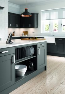 Anthracite shaker units create a modern twist in this traditional kitchen design. White worktops and chrome handles are displayed throughput the kitchen. This viewpoint shows us the features in the kitchen island. Smart plate storage and open shelving with cupboards either side. In the open shelving plates are on display on the top rack and the bottom shelf has 3 white bowls stacked and a black water jug. On top of the island there is a chopping boards and knife set. In the background, there is a Belfast sink in white and the kitchen windows allow light to flow in.