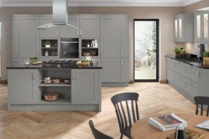 A shaker kitchen layout, using dust grey Olympia shaker doors. The kitchen has silver handles, a kitchen island and two runs of units - one tall unit run complete with single over and open shelving either side. The other run of units is to the right of the island, containing 6 kitchen drawers with a stainless steel sink and top in the middle on top of the granite worktop. there are 2 glass fronted cabinets either side of the sink with a window in the middle of the units directly above the sink. A wooden topped round kitchen table is blurred in focus, in front of the kitchen island with some books on top of it. The Kitchen has a wooden floor and the kitchen has cashmere painted walls.