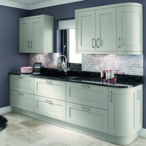 This is showing one top and bottom run of a kitchen design. There are 6 drawers and one curved unit along ht bottom run showcasing light grey shaker doors and units. A black granite worktop, matte black sink and chrome tap are placed on top of the counter in the middle of the run just under the kitchen window. The top run of units has 4 units with light grey shaker doors and with chrome handles. The following has cashmere slab tiles and dusty grey painted walls. There is an iPad on top of the counter and 4 bottles of rose wine. Hand creams and soaps are placed beside the sink. The wall between the top and bottom units is tiled with natural light stone.