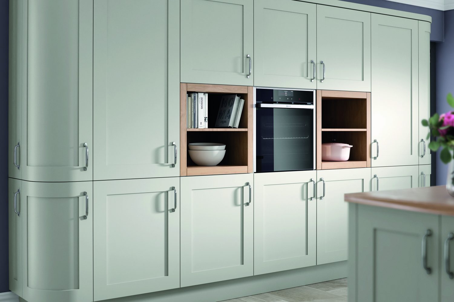 One wall of the Kitchen is showcased here, showcasing shaker light grey Olympia units. Tall larders with curved units are on the left, a singe over with open shelving either side are In the middle and one curved tall unit is on the other side. The walls are dusty grey with cashmere slabbed tiling on the floor. There are cook books, a large ivory casserole dish and 8 cook books on the open shelving either side of the oven. The worktop on the kitchen island which is only slightly in view is wooden