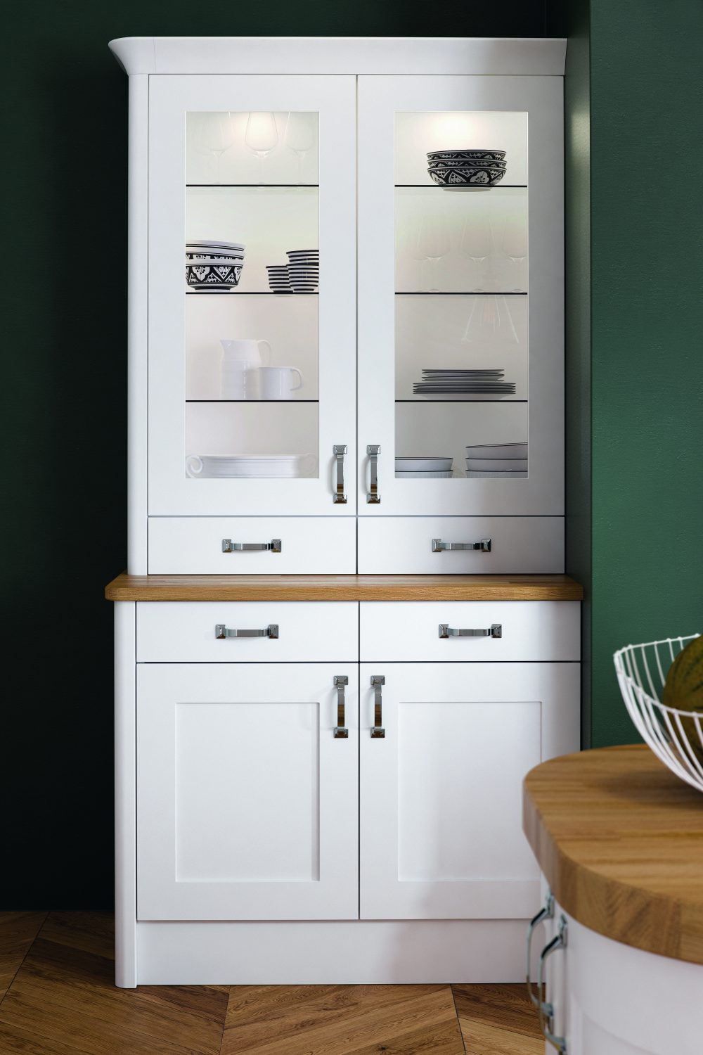 White Olympia shaker kitchen units are displayed in this dresser. The dresser has glass fronts on top, showcasing plates and bowls that are stored away neatly. The bottom half of the dresser has two top drawers and two cupboard doors underneath. The handles are chrome.