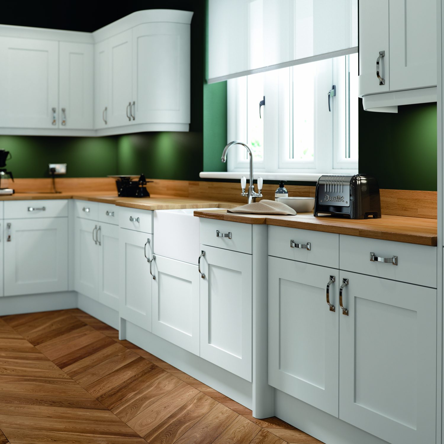 A beautifully traditional style with a modern twist, this kitchen design include Olympia white shaker doors and kitchen units. The walls are green and the floor is a lighter herringbone wood. The worktops are wooden with a toaster on display to the right of the sink. The sink is a white Belfast style sink, with chrome tap. The kitchen handles are chrome. The kitchen window is placed behind the sink and is letting light in with the white blinds half opened.